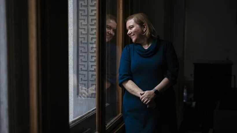 Dr. Allison Arwady stands smiling by tall windows in a teal dress, her reflection visible