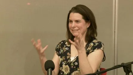 Anna Clark gestures with both hands while speaking at a microphone in a floral dress