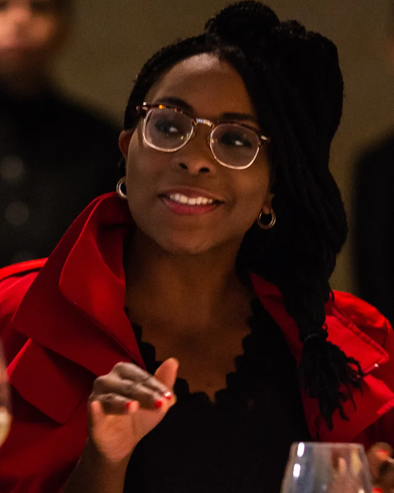 Belinda Archibong in a red blazer and glasses, mid-gesture while speaking at a dinner table