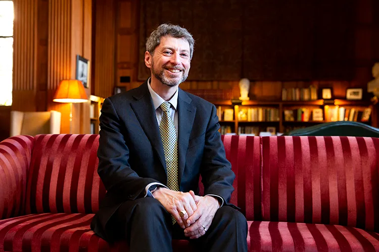 Benjamin Hermalin sitting on a red tufted velvet sofa inside a campus building