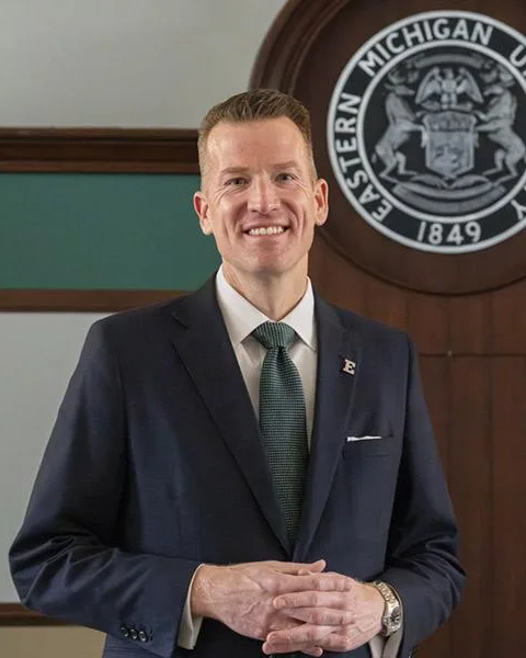 Brendan Kelly in a navy suit and green tie before the Eastern Michigan University seal