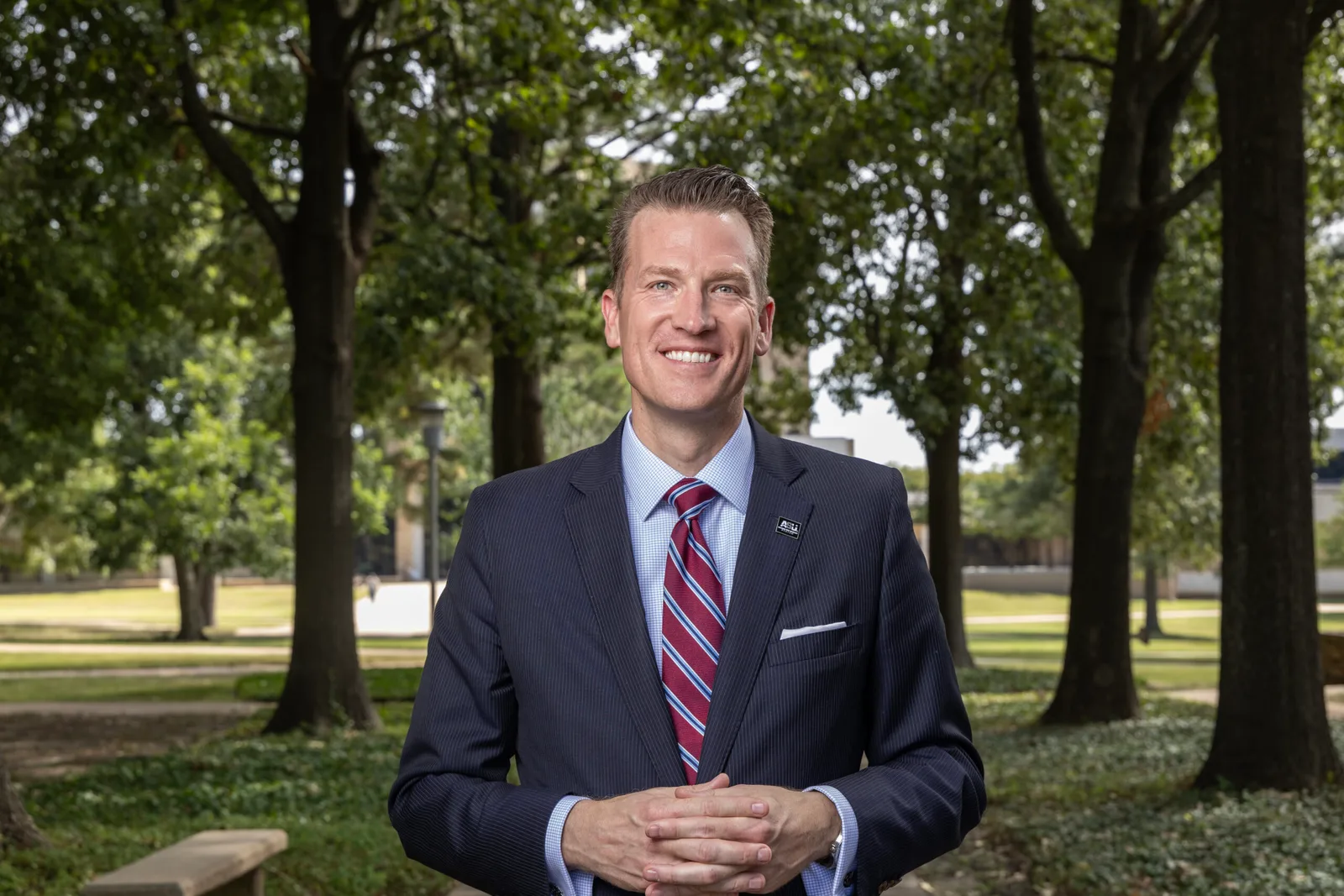 Brendan Kelly in a navy pinstripe suit and striped tie standing outdoors among campus trees