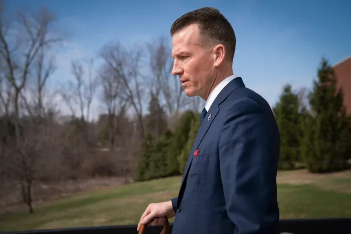 Brendan Kelly in profile wearing a navy suit outdoors, with bare trees and evergreens behind