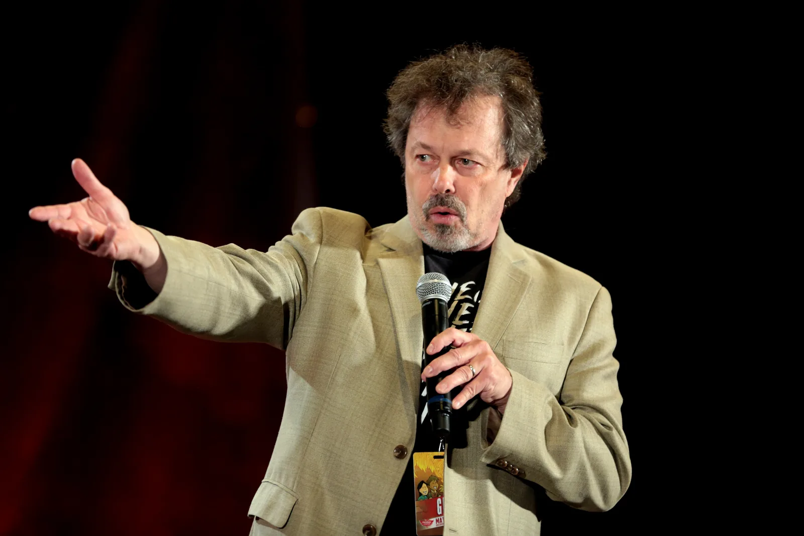 Curtis Armstrong in a tan blazer speaking into a microphone on stage, one arm stretched out to the side