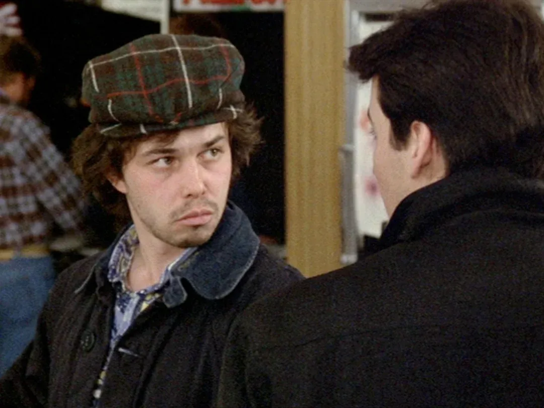 Curtis Armstrong in a plaid cap and dark coat glancing sideways at another man in Better Off Dead