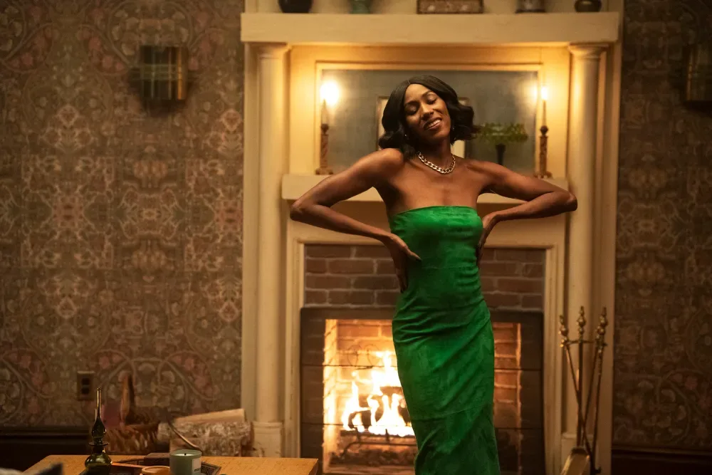 Diarra Kilpatrick in a green gown, laughing, in front of a lit fireplace, in a scene from Diarra from Detroit