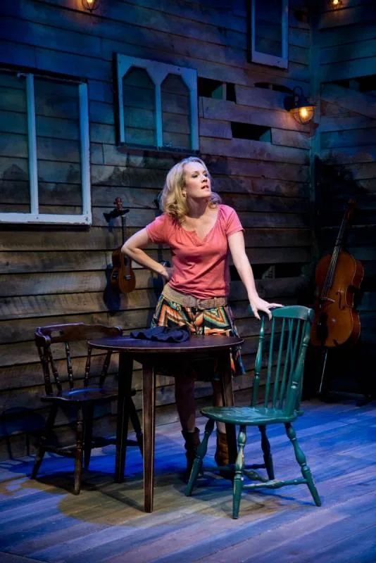 Erin Dilly stands beside a table and green chair on a rustic wooden stage set