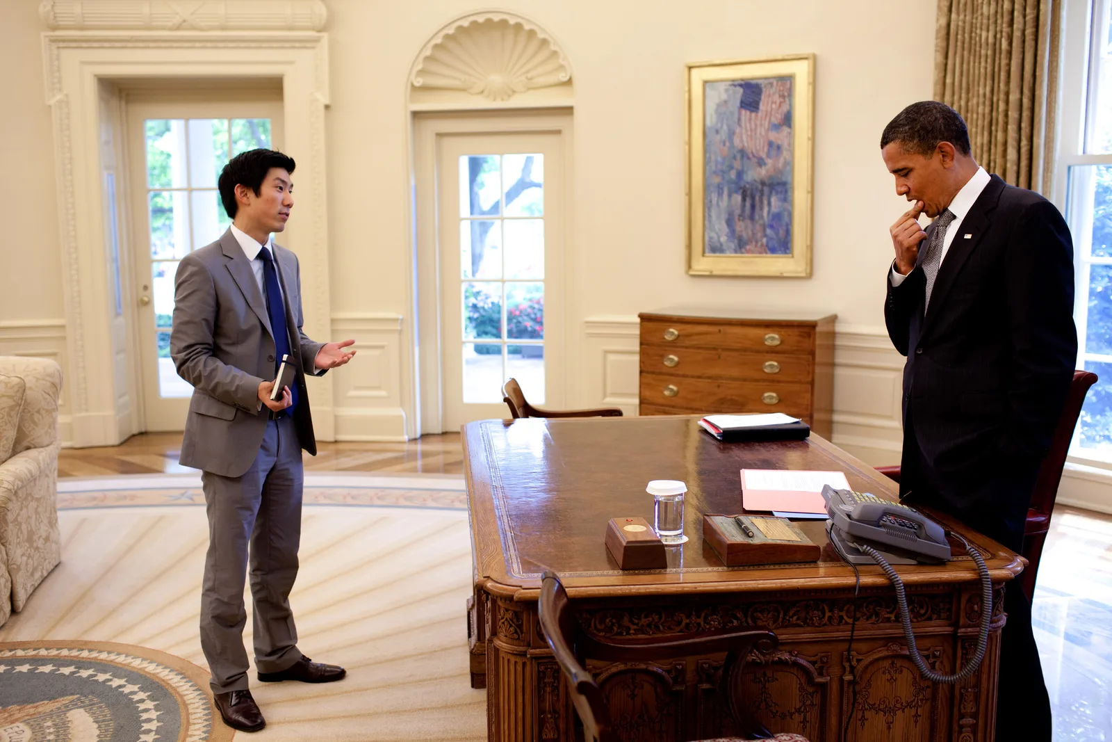 Eugene Kang standing in the Oval Office facing President Barack Obama, who is on the telephone at the Resolute Desk