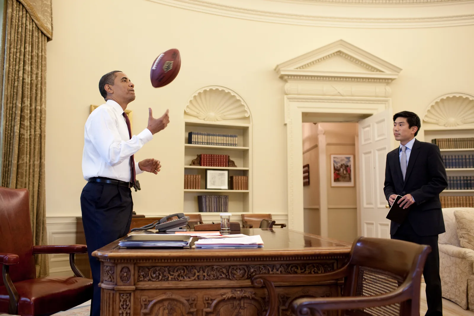President Barack Obama tossing a football across the Oval Office while Eugene Kang watches from the doorway