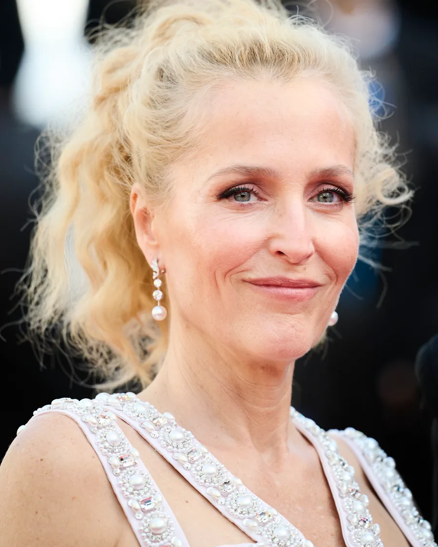 Gillian Anderson smiling on a red carpet, blonde hair up, pearl drop earrings and beaded gown
