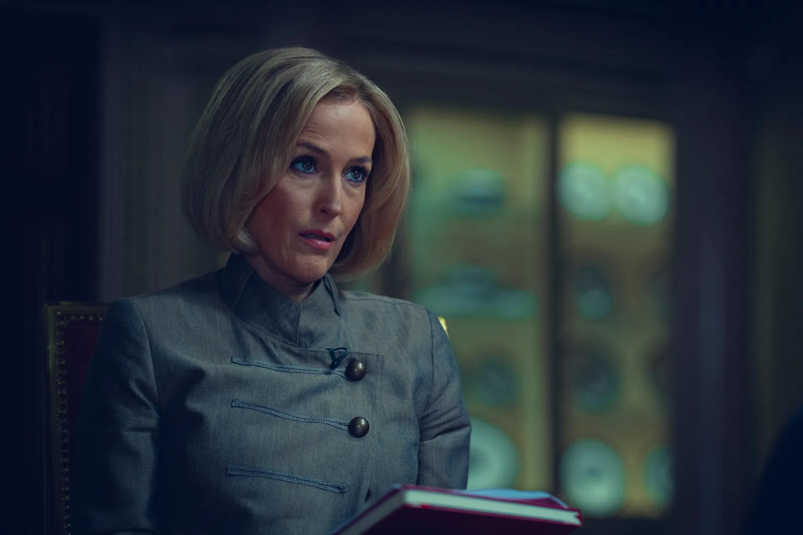 Gillian Anderson in a grey high-collared jacket seated in an interior, holding a red book