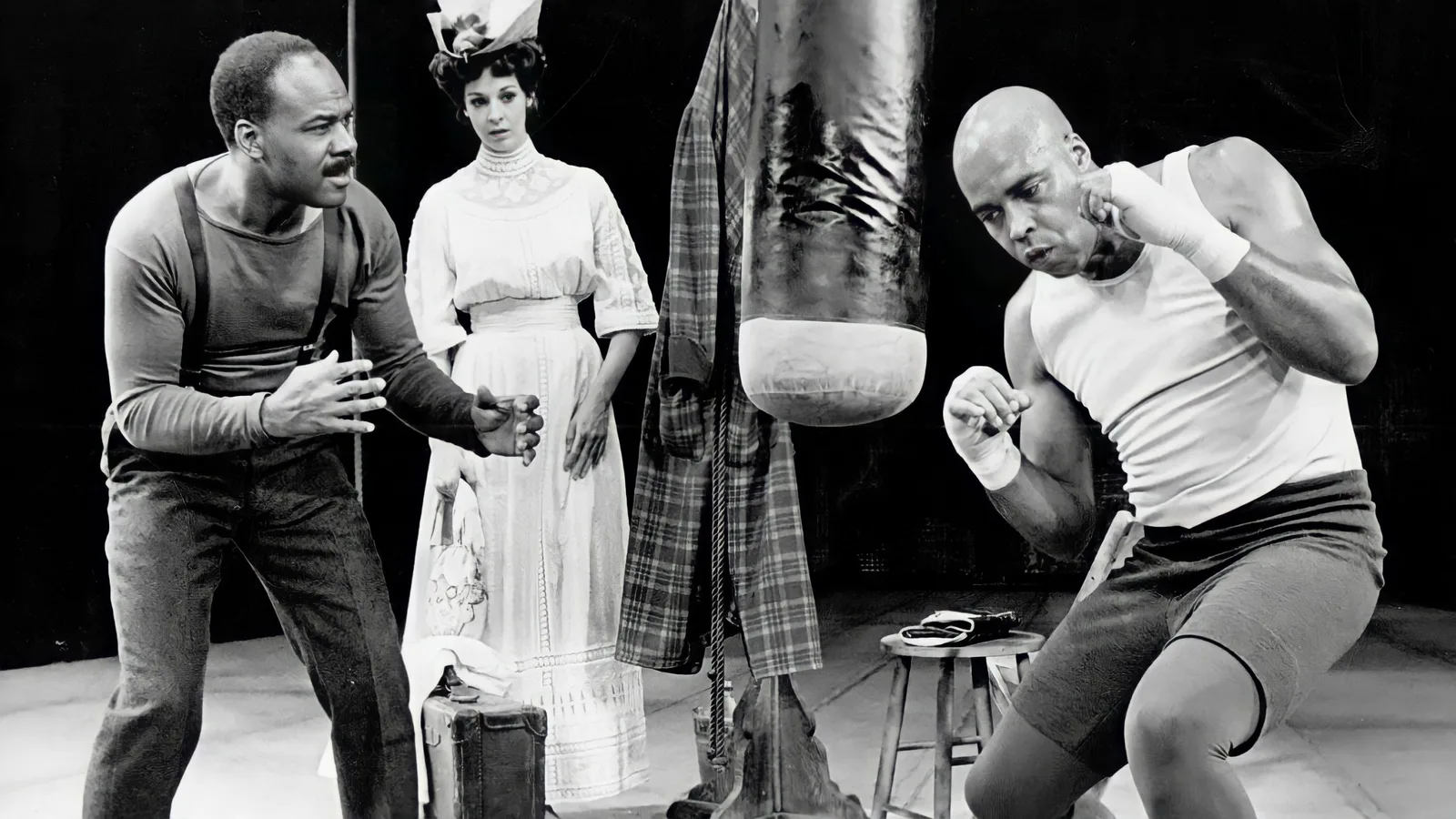 Black and white stage still: James Earl Jones in training, throwing a punch as two performers watch