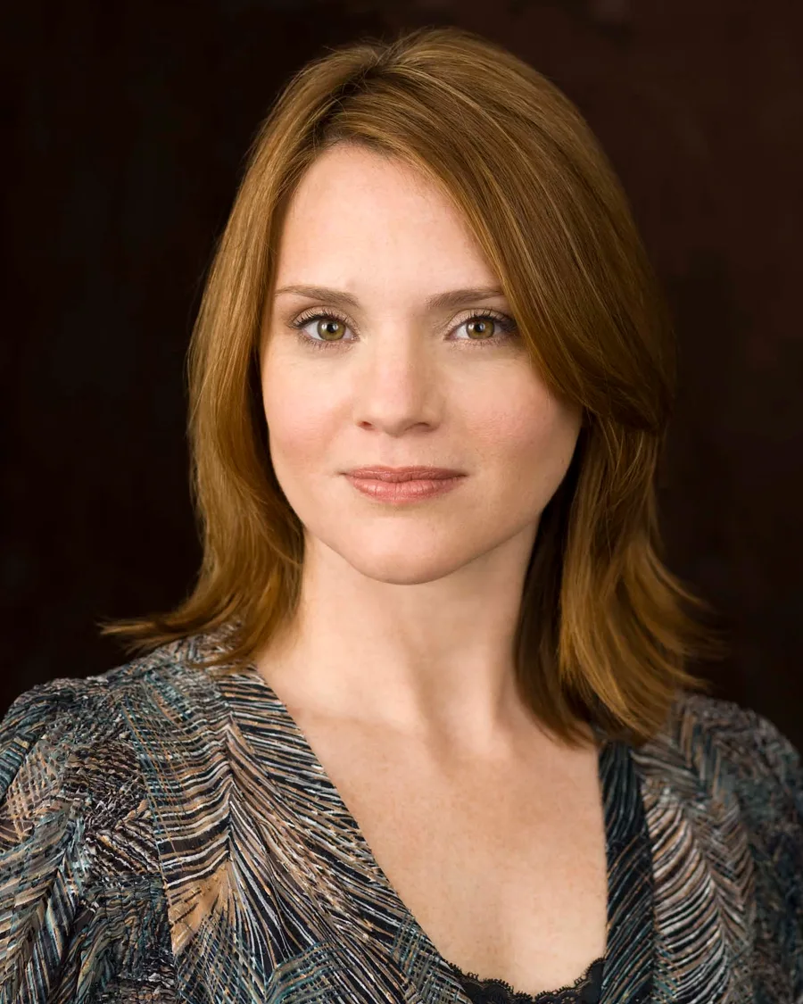 Jennifer Laura Thompson in a patterned top, auburn hair to her shoulders, against a dark background