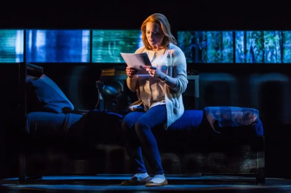 Jennifer Laura Thompson seated on a bed reading pages on a blue-lit stage set