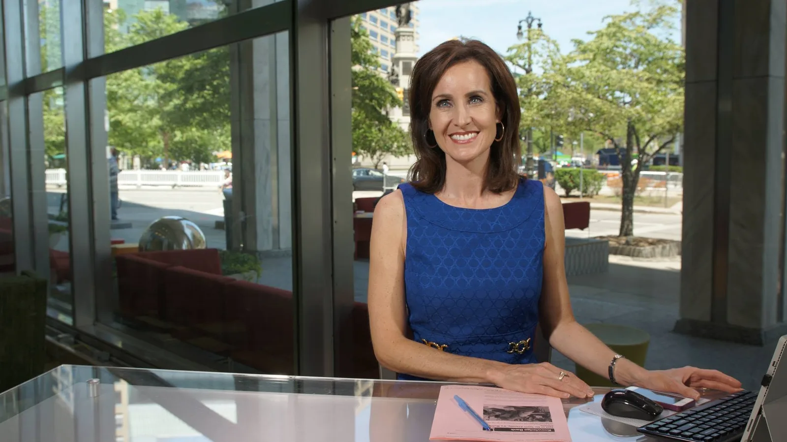 JoAnne Purtan smiles at a glass desk with keyboard and papers beside a downtown window