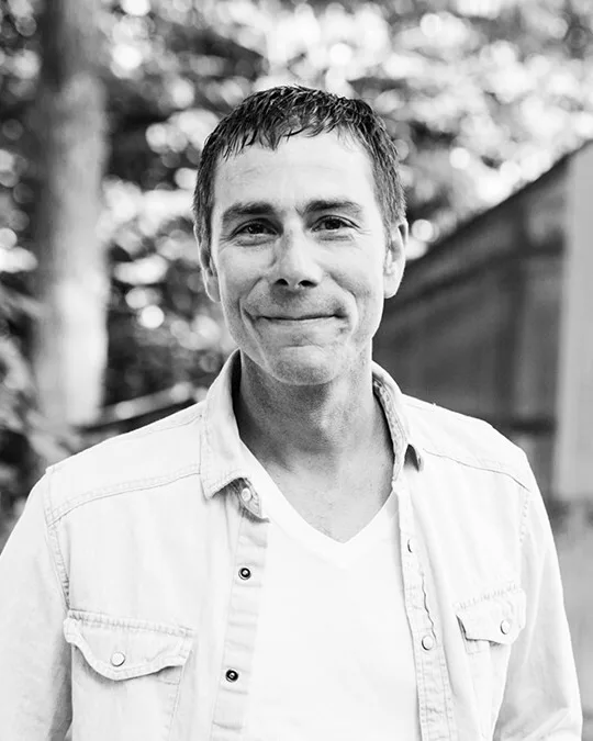 Black-and-white portrait of Joseph Beyer in an open work shirt outside a clapboard house