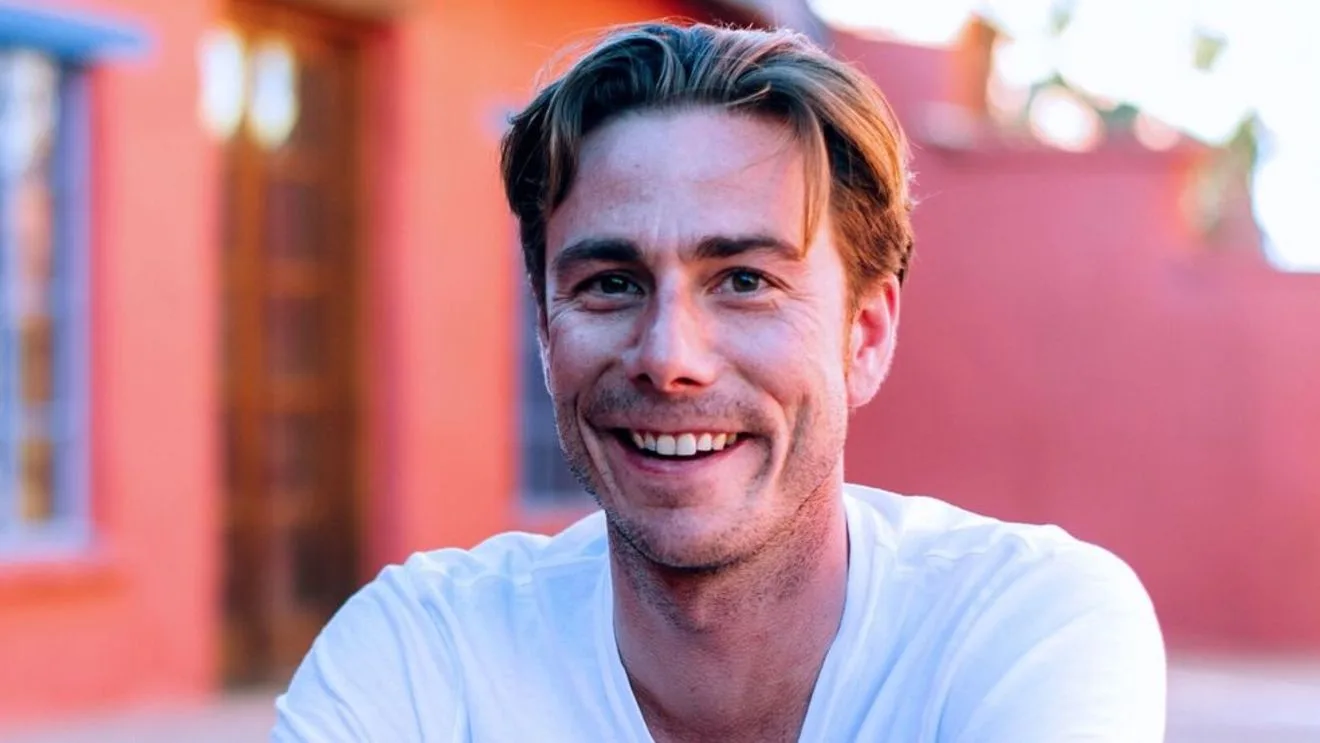 Joseph Beyer smiling broadly in a white t-shirt against a coral-painted wall in warm light