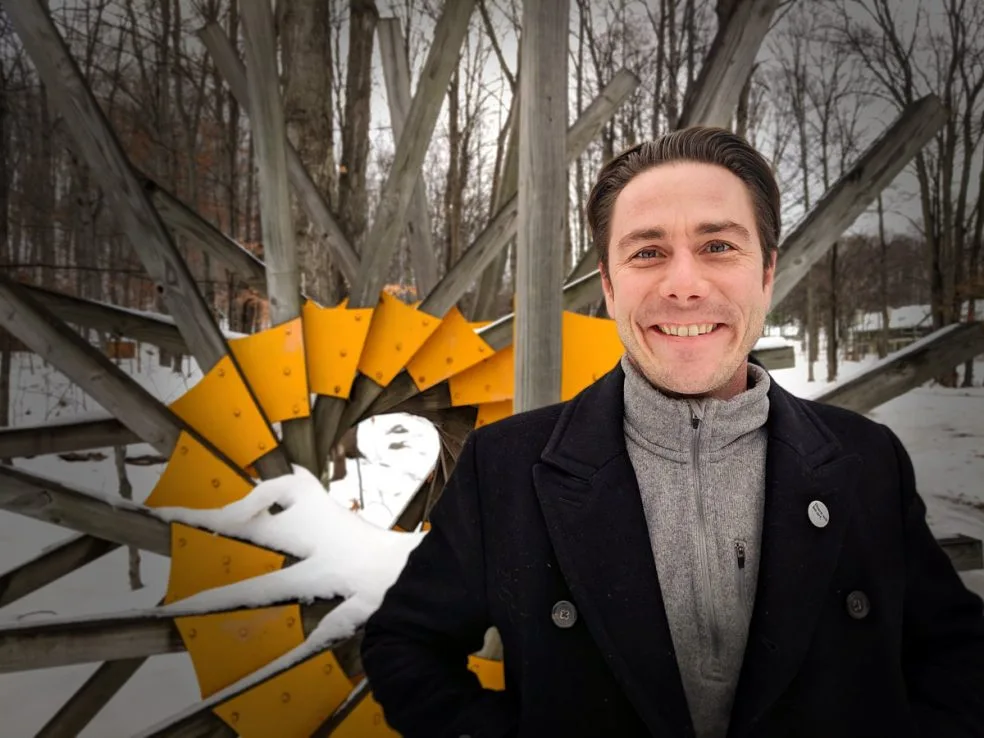 Joseph Beyer in a black overcoat smiling beside a yellow and wood sculpture in snowy woods