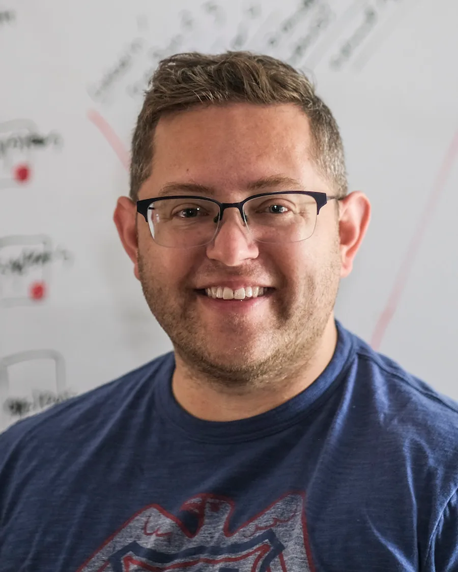 Joshua Steinman smiling in a blue T-shirt in front of a whiteboard covered in hand-drawn system diagrams