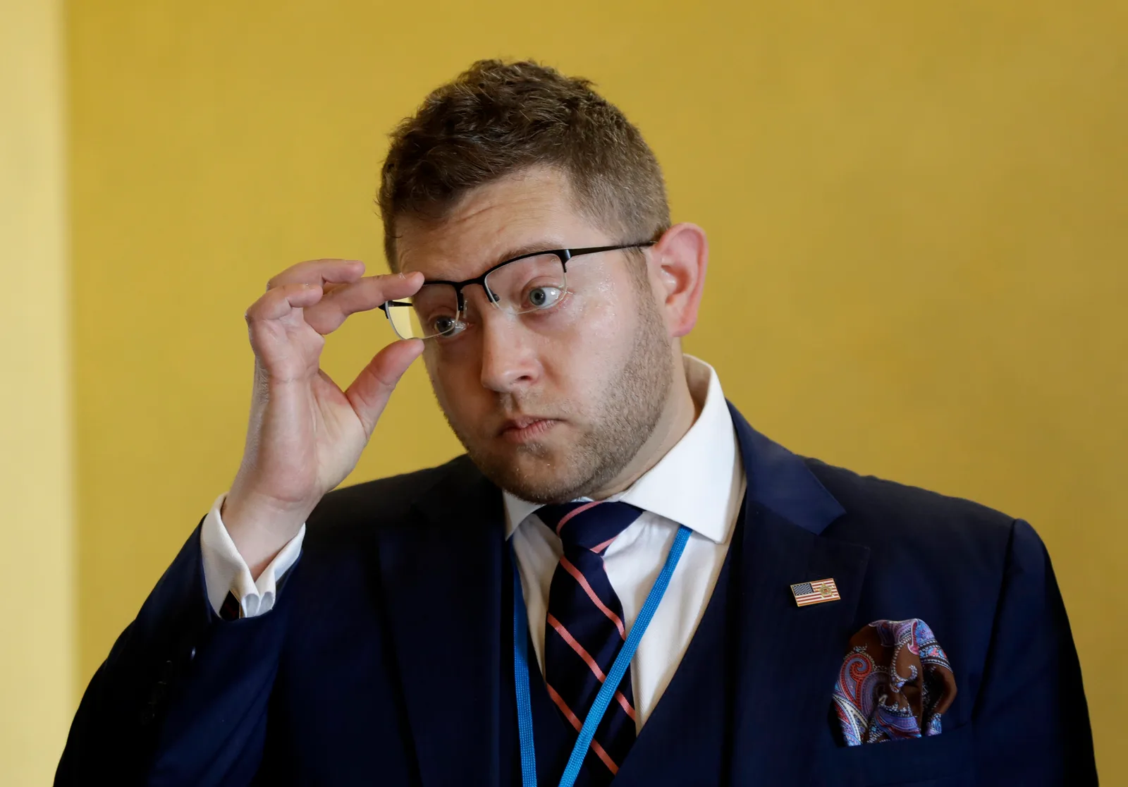 Joshua Steinman in a navy three-piece suit and lanyard, lifting his glasses with one hand