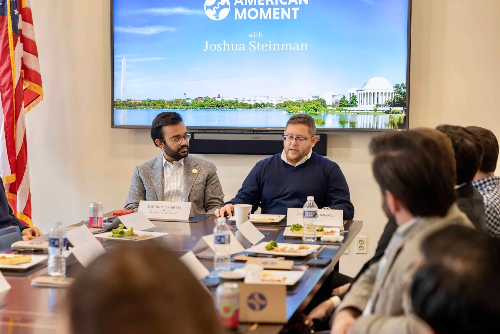 Joshua Steinman speaking at a conference table beside Saurabh Sharma, a screen behind them reading American Moment