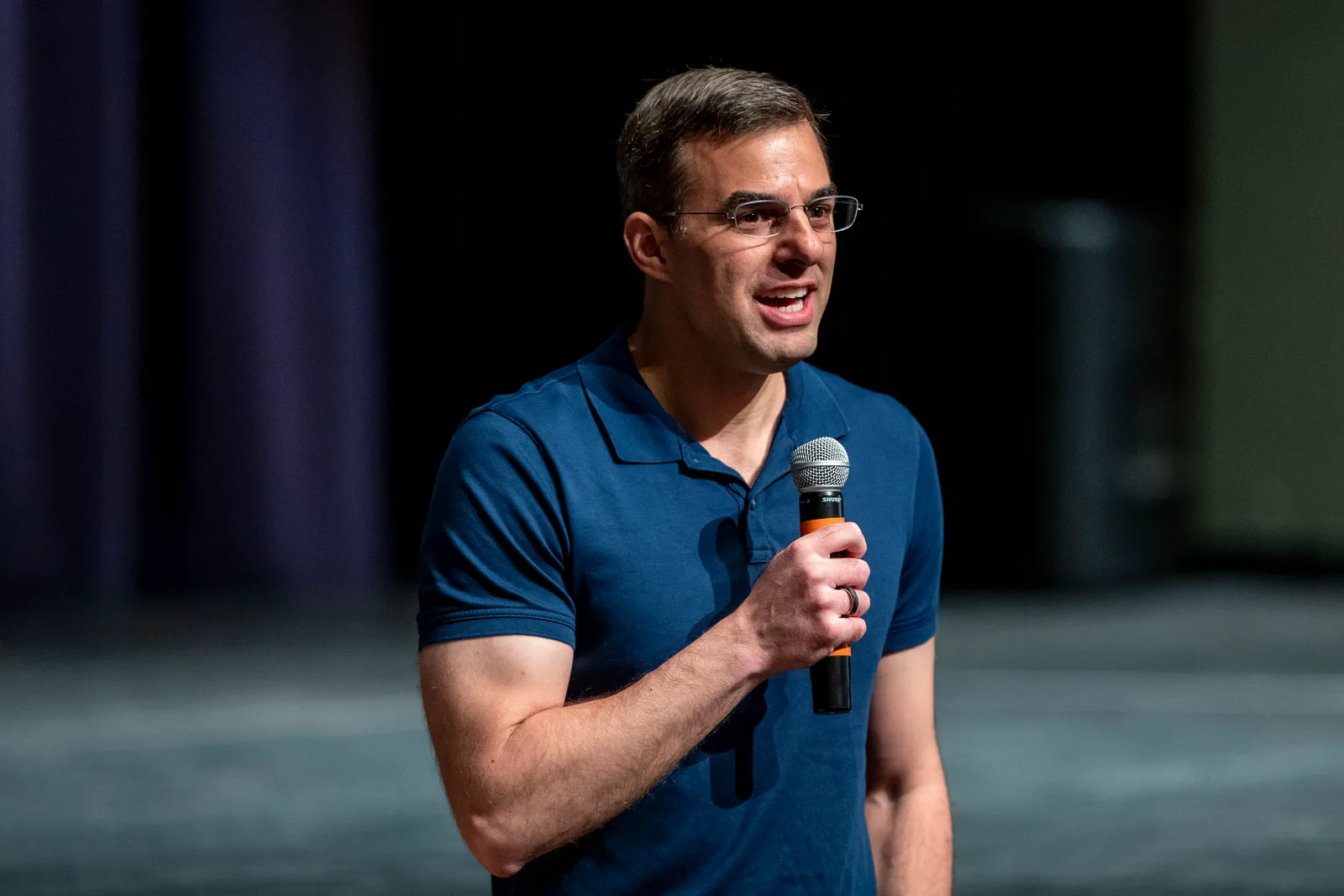 Justin Amash in a blue polo shirt speaks into a handheld microphone on a dark stage