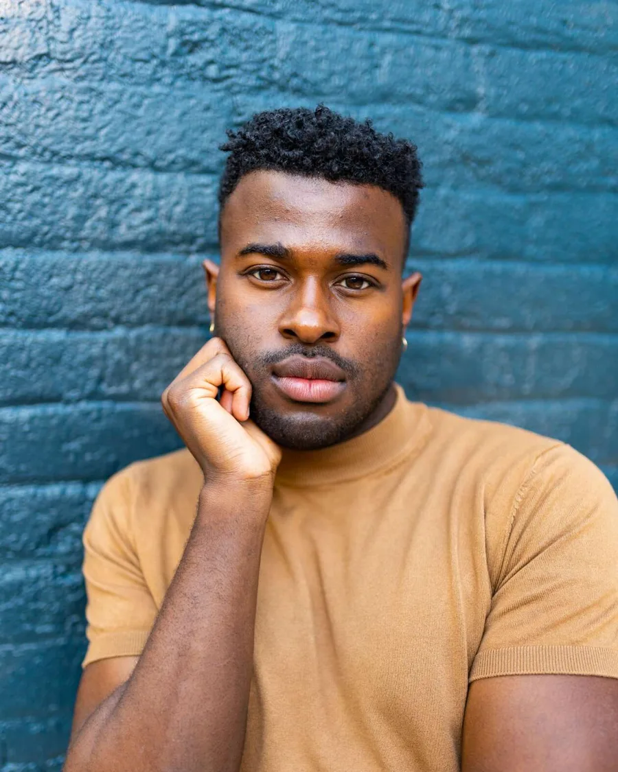 Lee Alexander rests his chin on his hand in a tan knit top against a blue painted brick wall