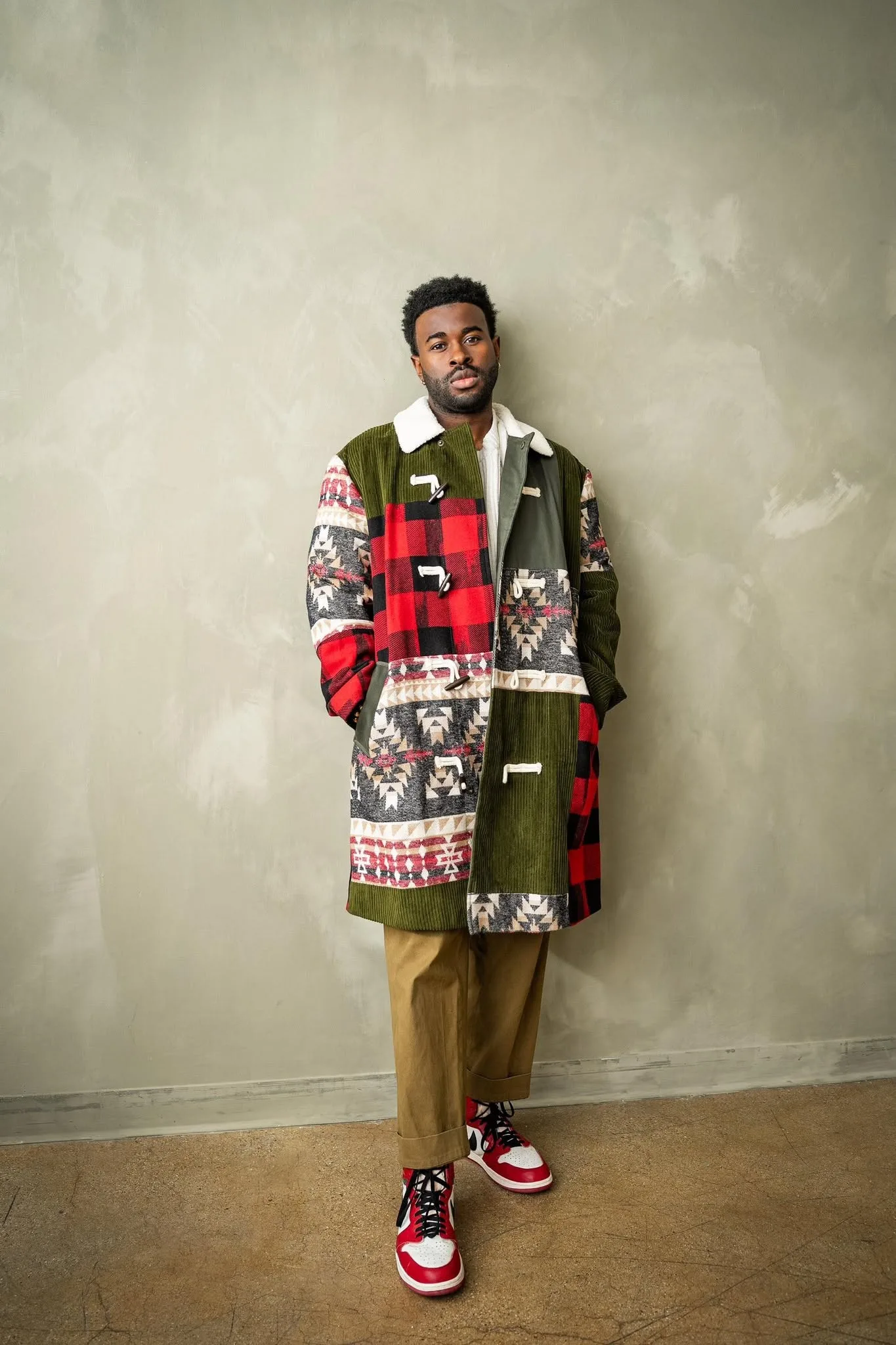 Lee Alexander stands full length in a patchwork corduroy and plaid coat against a plaster wall