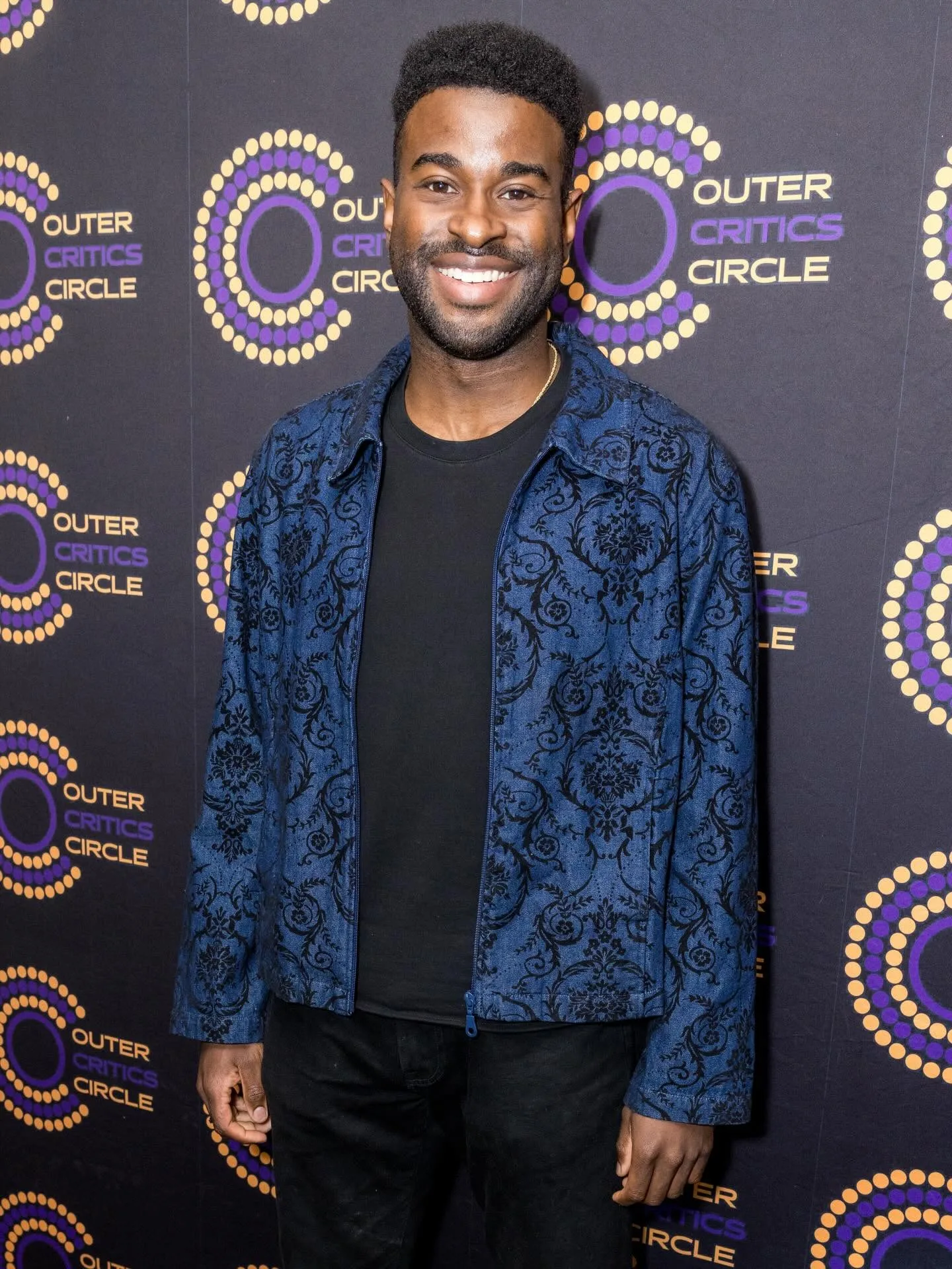 Lee Alexander in a patterned blue jacket at an Outer Critics Circle step-and-repeat wall
