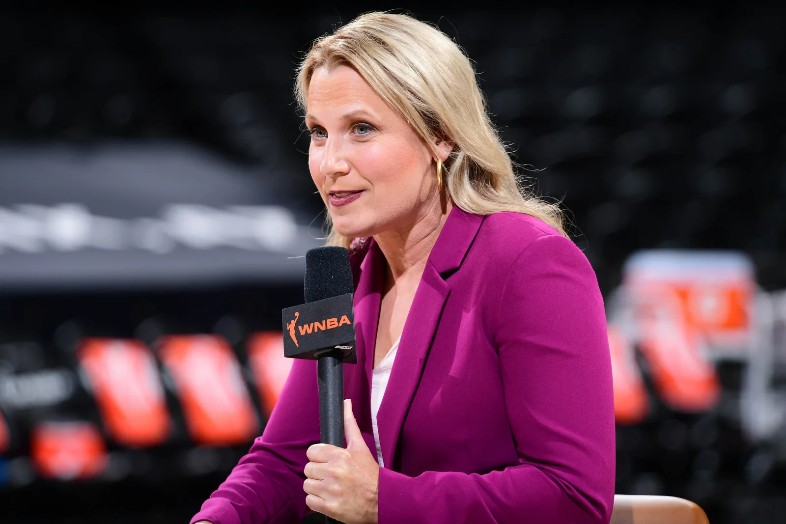 Lisa Byington in a magenta blazer holding a WNBA-branded microphone inside an empty basketball arena