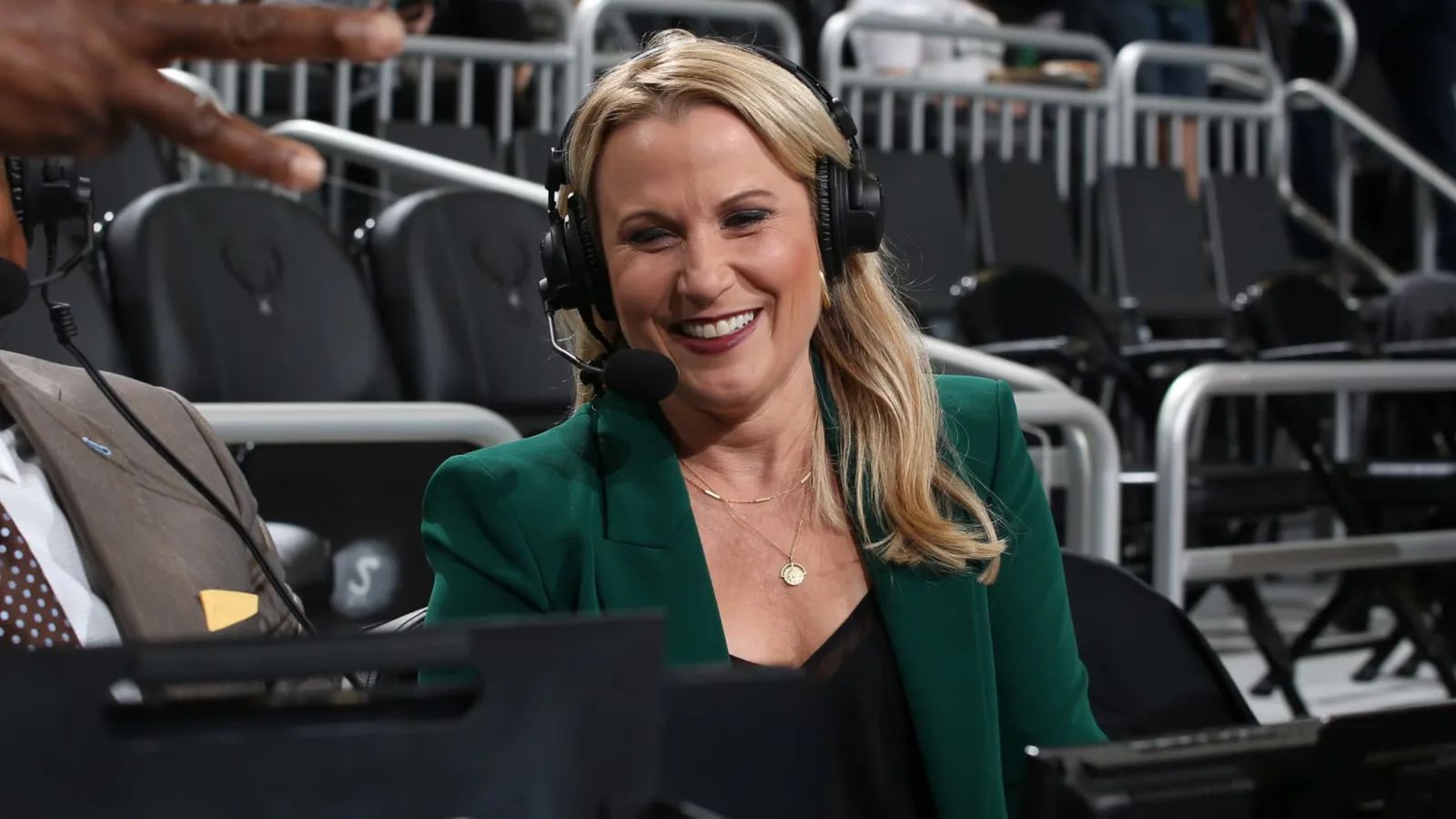 Lisa Byington smiling at a courtside broadcast table wearing a headset microphone and green blazer