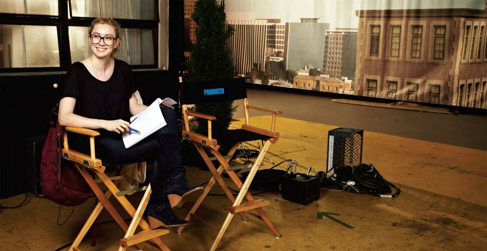 Liz Meriwether seated in a director's chair marked PRODUCER on a studio backlot, script in hand, a city skyline backdrop behind her