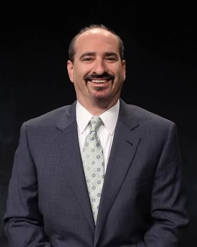 Mario Impemba in a gray suit and patterned tie smiles against a black background