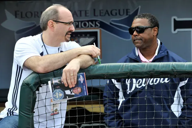 Mario Impemba with a notebook leaning on a dugout rail, talking with Rod Allen in Detroit gear