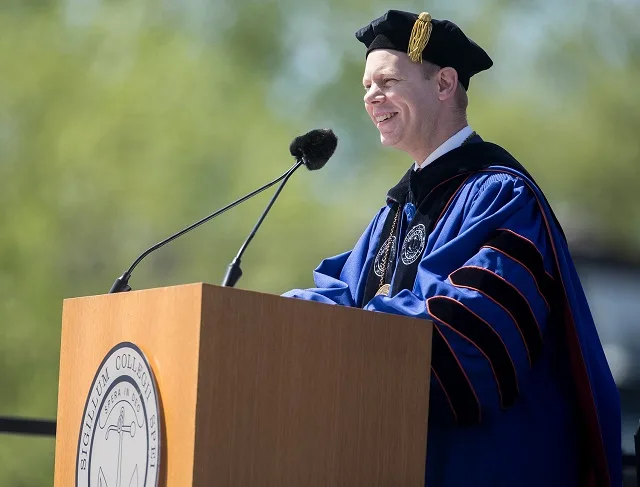 Matthew Scogin in blue academic regalia and mortarboard speaks at an outdoor lectern