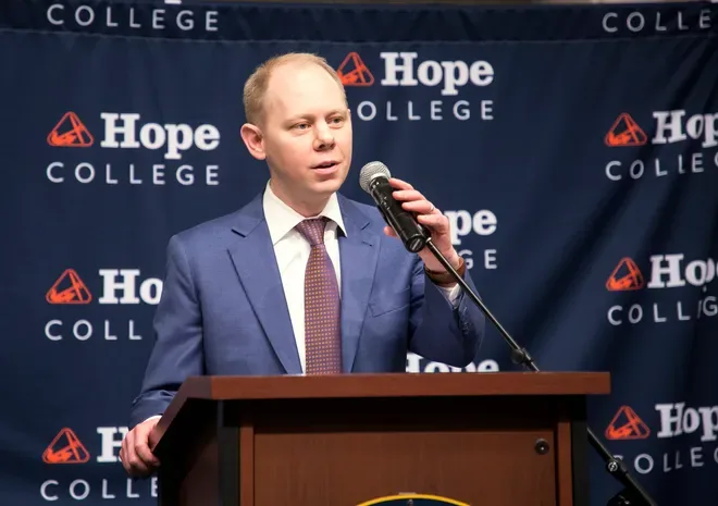 Matthew Scogin speaks into a microphone at a lectern before a Hope College backdrop