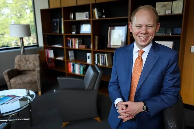 Matthew Scogin in a blue check suit and orange tie stands smiling in an office with bookshelves
