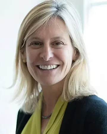 Molly McUsic smiles in a black jacket over a yellow blouse against a white background
