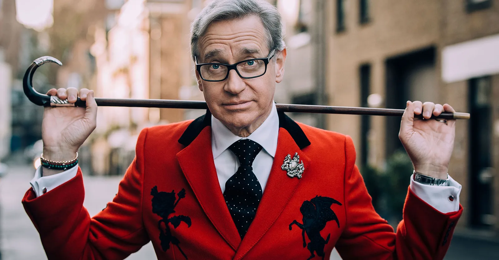 Paul Feig in a red appliqued jacket holds a walking cane across his shoulders on a street