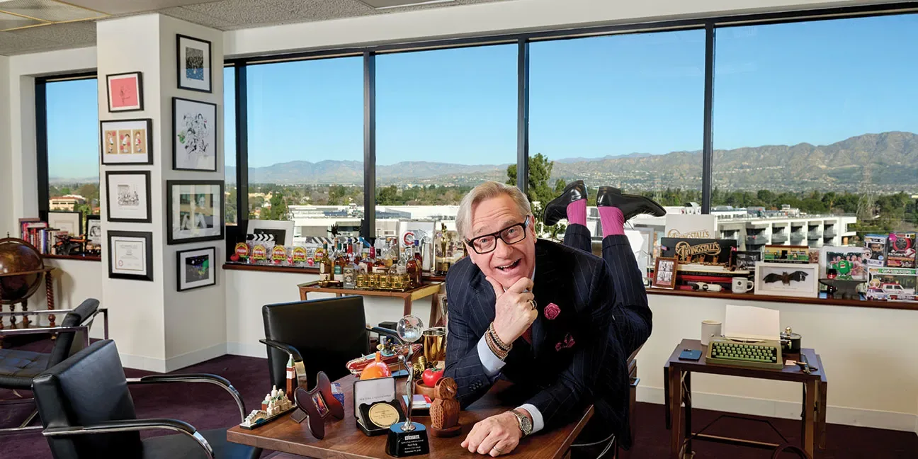 Paul Feig sprawls across a desk with his feet in the air in an office full of memorabilia