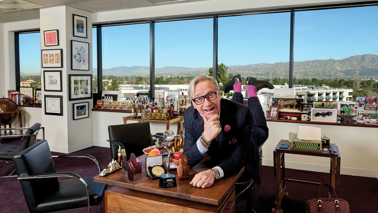 Paul Feig sprawls across a desk with his feet in the air in an office full of memorabilia