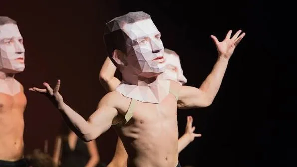 Shirtless performers in angular paper face masks pose with raised arms on a dark stage