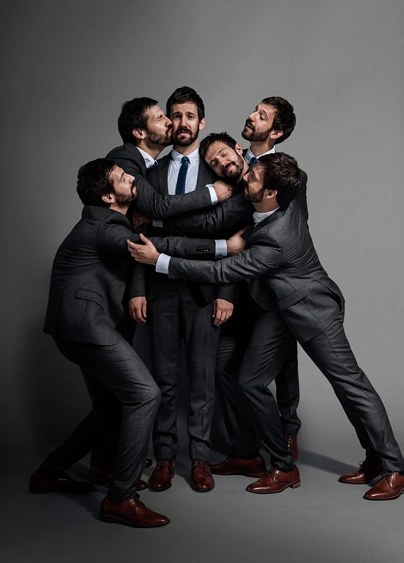 Five men in matching grey suits cluster together, four of them leaning on the one standing in the centre