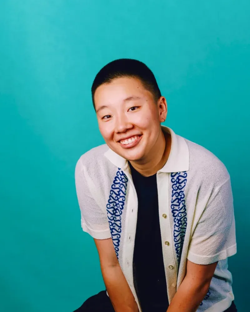 Sabrina Wu smiling, seated on a stool in a white patterned shirt against a teal backdrop