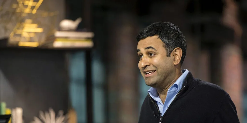 Sumir Meghani gesturing while speaking during a television interview, mugs on the counter in front of him