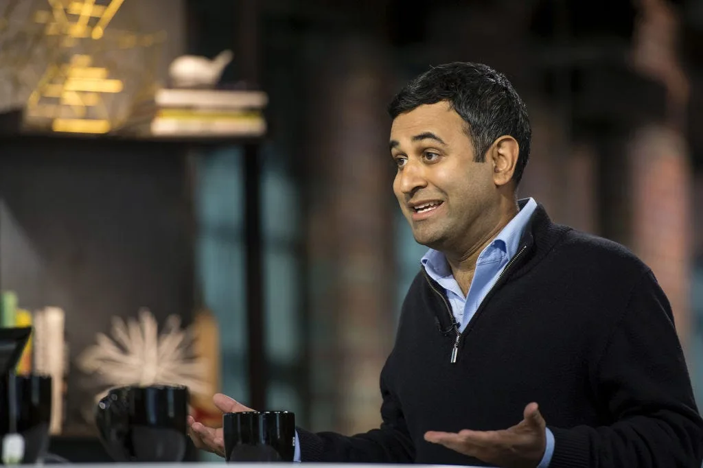 Sumir Meghani gesturing while speaking during a television interview, mugs on the counter in front of him