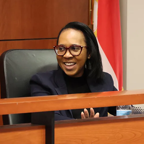 Judge Tiffany P. Cunningham in burgundy glasses and a navy blazer, seated at the bench with a flag behind her