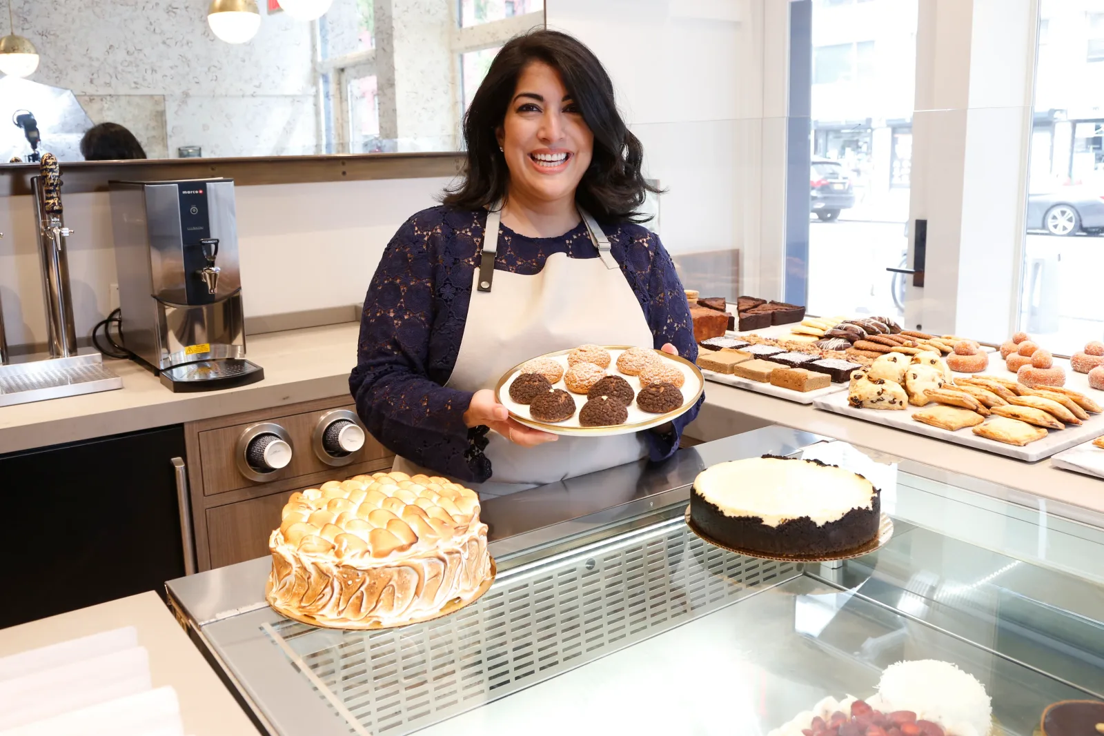 Umber Ahmad smiling behind a bakery counter, holding a plate of cookies, pastries and cakes displayed around her