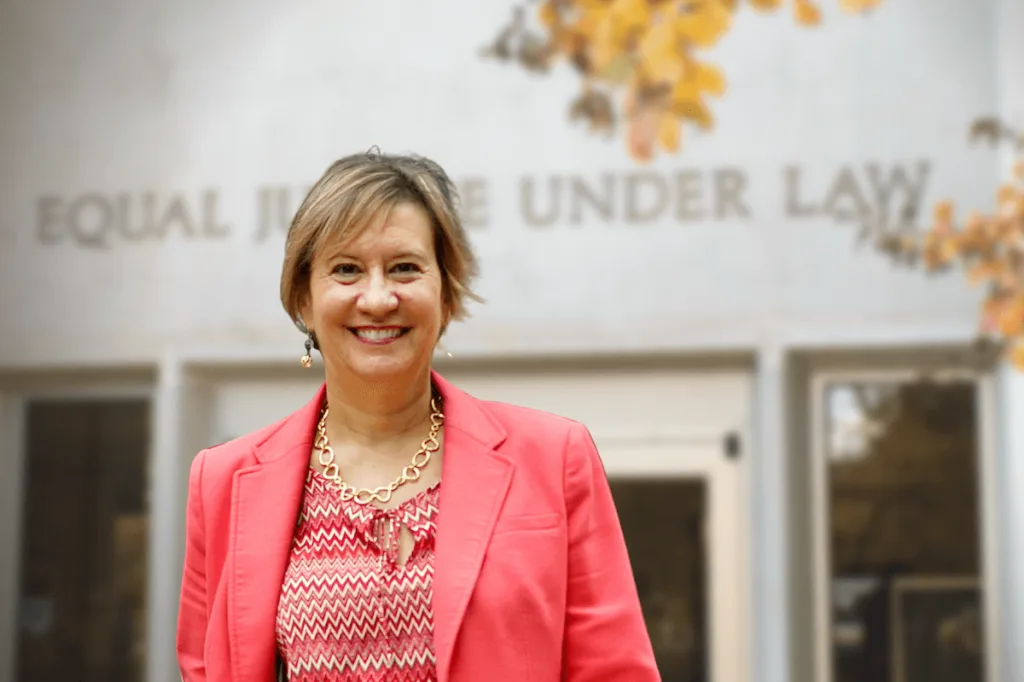 Valorie Vojdik smiling in a coral blazer in front of the College of Law building, with the words Equal Justice Under Law carved above her