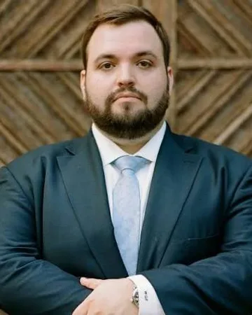 Wade Fink in a dark suit and light blue tie, arms folded, in front of a wooden door